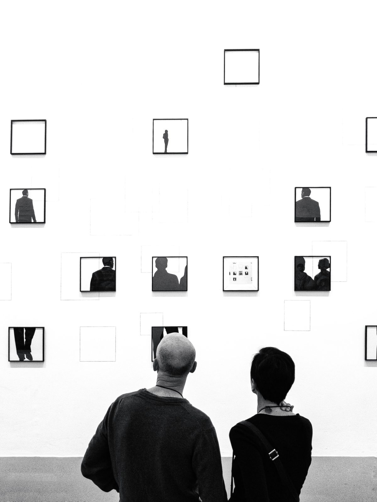 Deux visiteurs de dos regardent un mur blanc où sont accrochées de petites œuvres encadrées