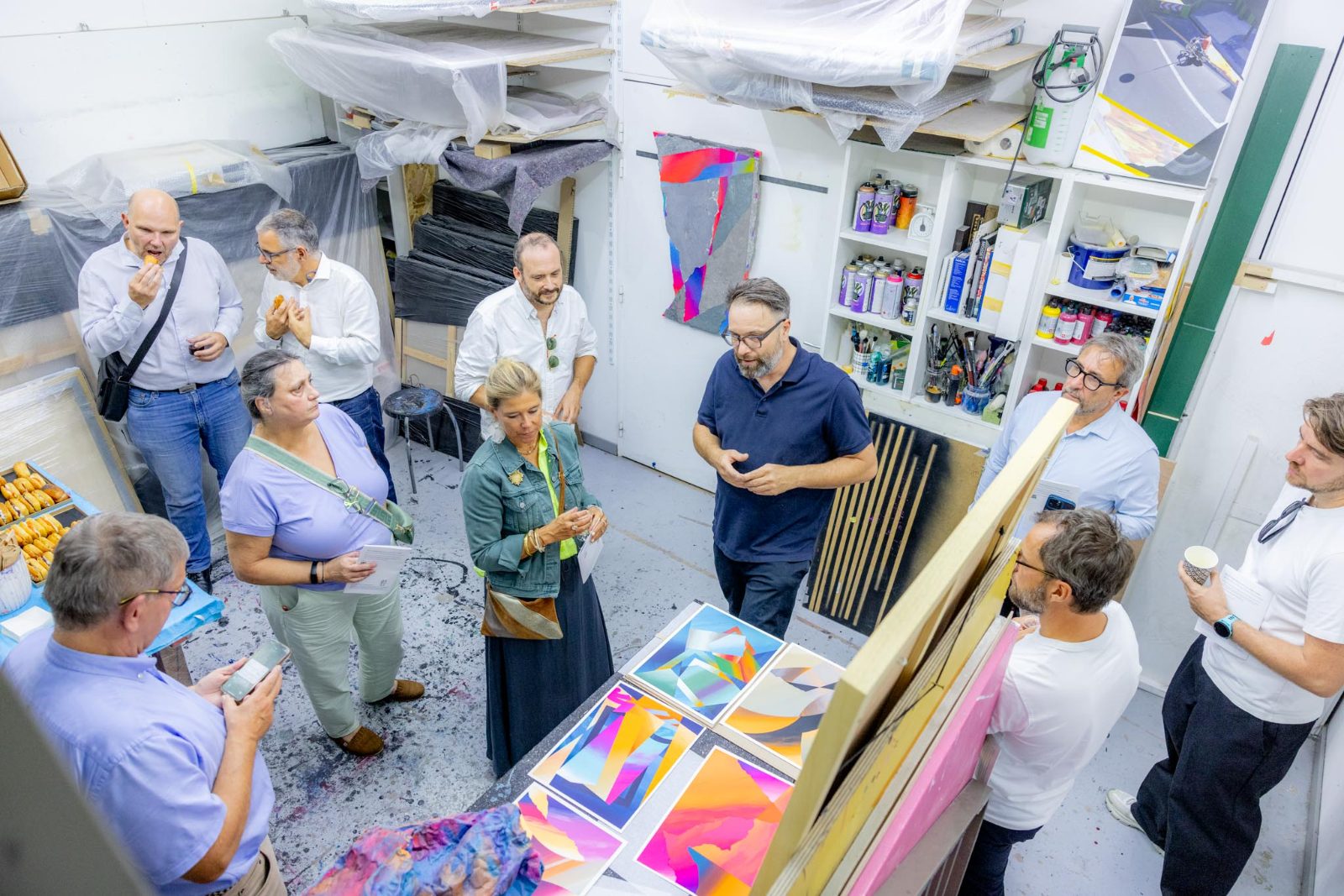 Vue en plongée d'un artiste s'adressant à une dizaine de participants réunis en cercle dans son atelier, entre œuvres et grandes toiles