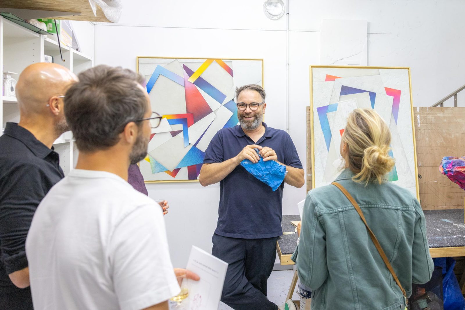 Dans un atelier, un artiste présente une pièce de papier froissé coloré à un petit groupe réuni devant de grandes toiles géométriques