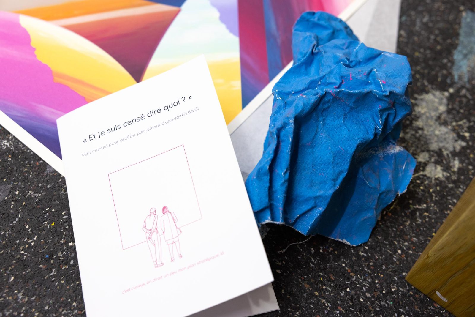 Le livret « Et je suis censé dire quoi ? » posé sur des estampes colorées, à côté d'une sculpture de papier bleu