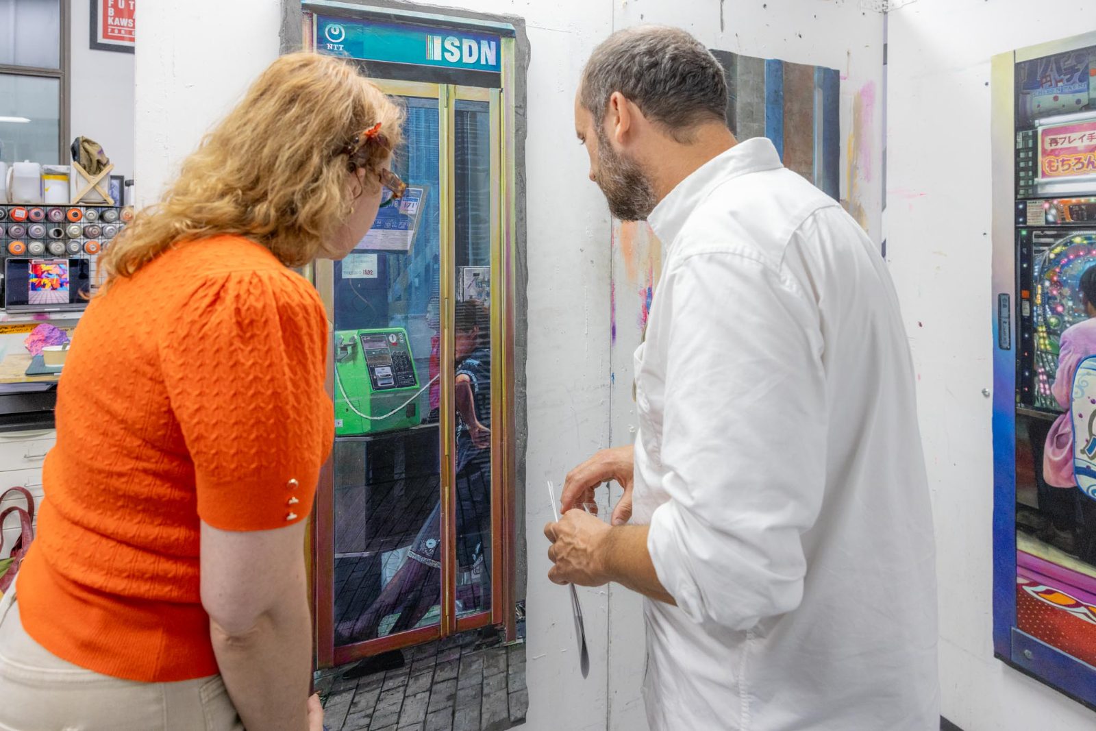 Deux visiteurs penchés vers une peinture représentant une cabine téléphonique, observant le détail de la matière