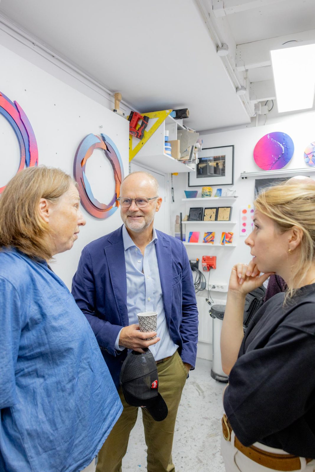Trois personnes en conversation détendue dans un atelier, devant des œuvres circulaires accrochées au mur